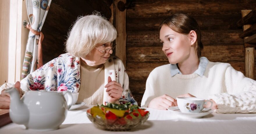 Older grandmother and teenage granddaughter talking over tea at a kitchen table, showing warm grandparent-grandchild bonding, family communication, and intergenerational connection