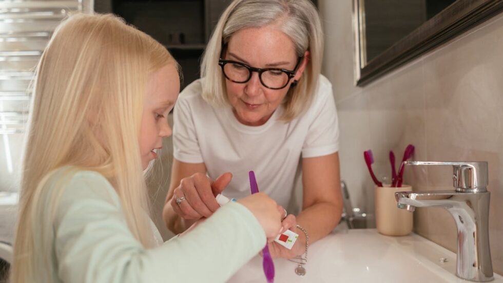 Grandmother helping her granddaughter brush her teeth at the bathroom sink, showing grandparent care, daily routines, and family bonding