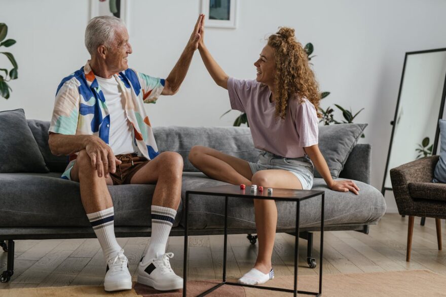 Older grandfather and young woman high-fiving on a sofa during a board game, representing grandparent-grandchild bonding, family connection, and intergenerational fun