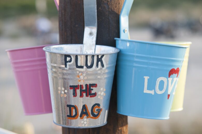 Colorful hanging metal buckets with painted words, including "PLUK THE DAG" and "LOVE," displayed outdoors on a post.