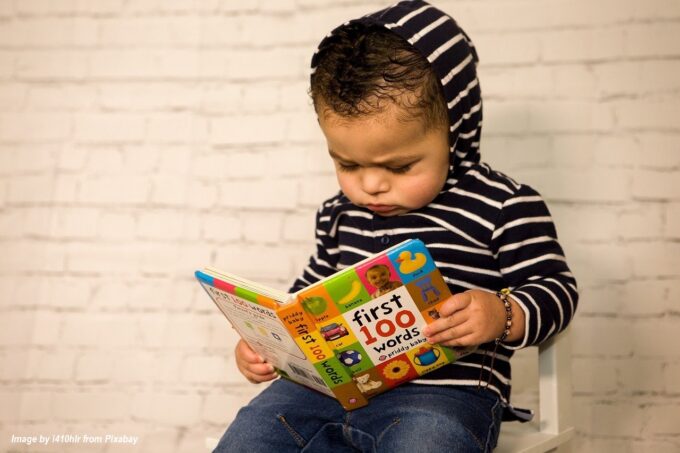 Toddler in a striped hoodie sitting on a chair and looking at a colorful early-learning board book, encouraging literacy and family learning on CaringGrandparents.com.