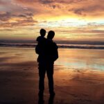 Grandparent carrying a grandchild along the beach at sunset, capturing a peaceful moment of intergenerational love and family bonding.
