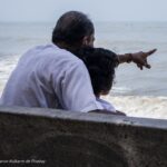 Grandparent and grandchild sitting together by the ocean, pointing toward the waves and enjoying a peaceful moment of intergenerational family bonding.