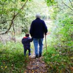 Grandparent holding a young grandchild’s hand while walking along a wooded trail, enjoying outdoor family time and intergenerational bonding.