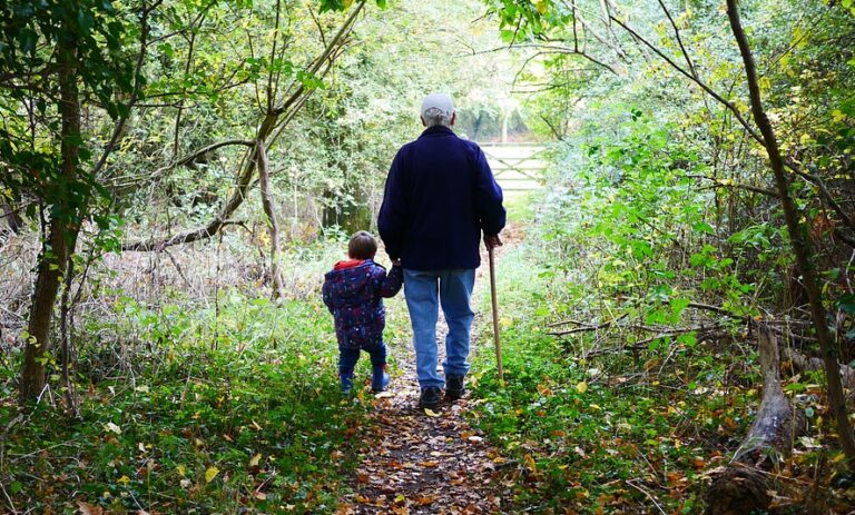 Grandparent holding a young grandchild’s hand while walking along a wooded trail, enjoying outdoor family time and intergenerational bonding.