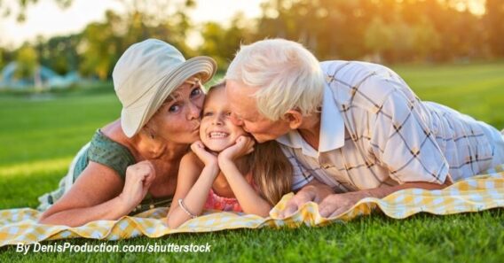 Grandparents kissing their smiling granddaughter while relaxing together on a picnic blanket in a sunny park.