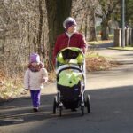 Grandparent walking with a young granddaughter and pushing a baby stroller along a neighborhood path, illustrating multigenerational family caregiving and outdoor bonding.