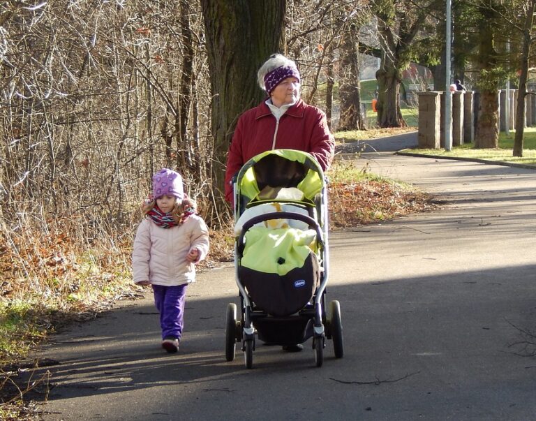 Grandparent walking with a young granddaughter and pushing a baby stroller along a neighborhood path, illustrating multigenerational family caregiving and outdoor bonding.