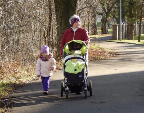 Grandparent walking with a young granddaughter and pushing a baby stroller along a neighborhood path, illustrating multigenerational family caregiving and outdoor bonding.