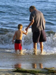 Grandfather holding his grandson’s hand as they play together in the ocean waves, enjoying quality intergenerational family time.