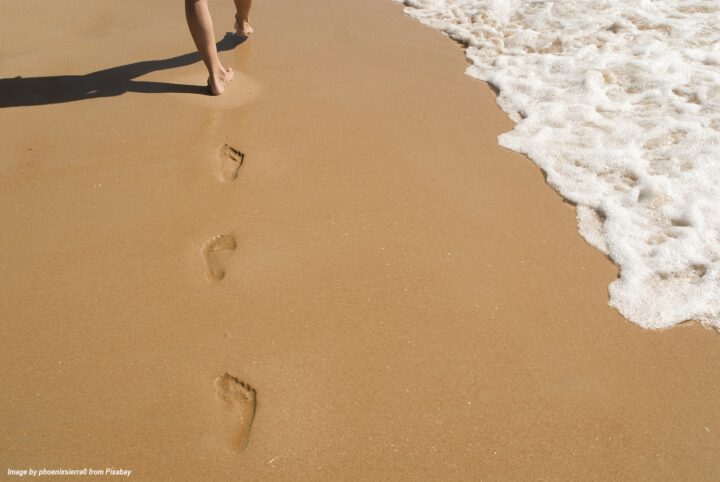 Bare feet leaving footprints in wet sand as a person walks along a sunny beach beside the ocean waves