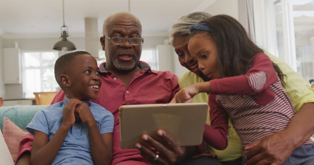Grandparents and grandchildren using a tablet together on a couch, showing multigenerational family bonding, shared learning, and quality grandparenting time.