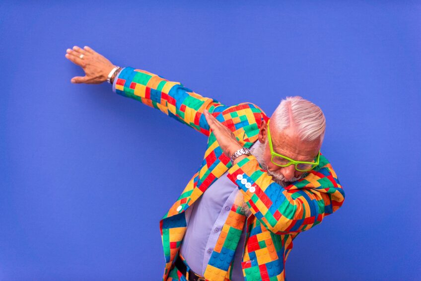 Senior man in a colorful patchwork jacket dabbing against a blue background, a playful multigenerational grandparenting lifestyle image.