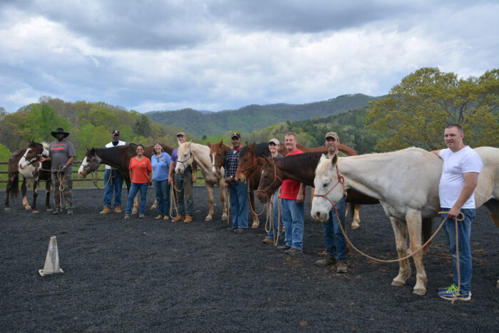 Group of people standing with horses in an outdoor riding arena, representing a multigenerational family activity and grandparenting connection.