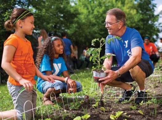 Older man gardening with two children outdoors, showing a hands-on grandparenting moment of teaching and family connection.