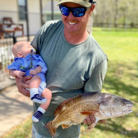 Man smiling while holding a baby and a large fish outdoors, a joyful grandparenting family moment.