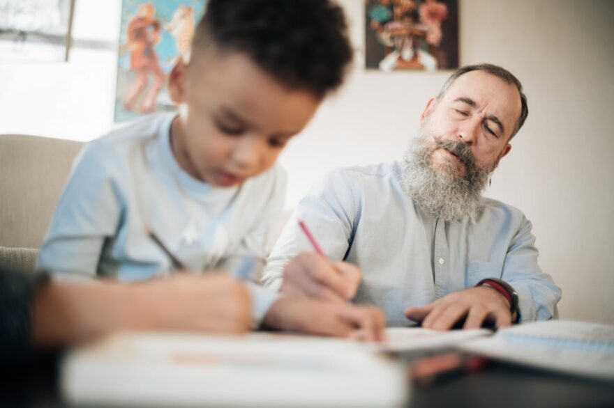 Grandfather helping a young child draw at a table, showing intergenerational learning, family bonding, and grandparenting
