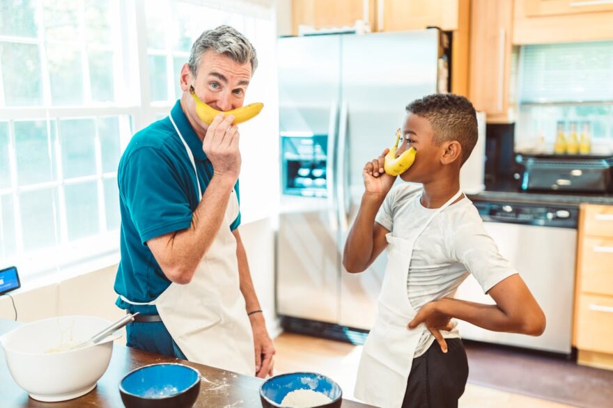 Grandfather and grandson in a kitchen pretending to use bananas as phones, showing playful grandparenting, intergenerational bonding, and family fun