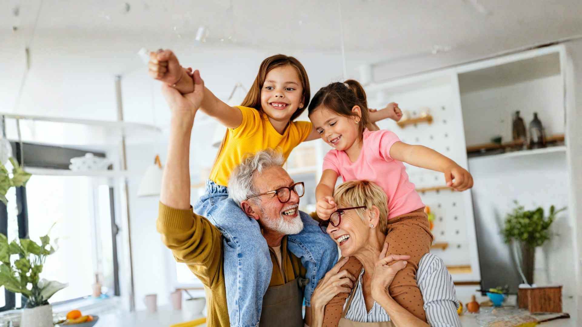 Grandparents and grandchildren laughing and playing together in a kitchen.
