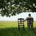 A man seated on a chair under a tree with an empty seat beside him.